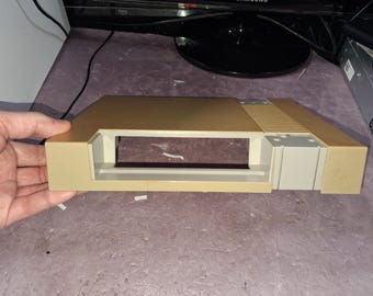 May include: A beige plastic top shell for a video game console featuring a rectangular opening for a cartridge slot and a series of horizontal ventilation slats on the right side. The casing has a smooth finish and a light tan color.