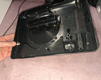 May include: A black plastic electronic device housing with an open interior, featuring a circular central cavity, a rectangular component mount, internal screw posts, and various molded structural ribs and connection points.