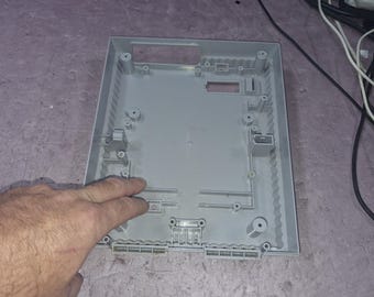 May include: A light gray plastic rectangular video game console shell casing featuring an open interior with molded screw posts, internal support structures, and rectangular cutouts for ports and connectors along the edges.