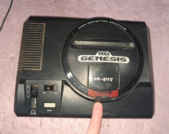 May include: A black Sega Genesis video game console featuring a cartridge slot, a volume slider, a reset button, and an on-off switch. The top circular panel displays the text High Definition Graphics, Sega Genesis, and 16-Bit in white lettering.