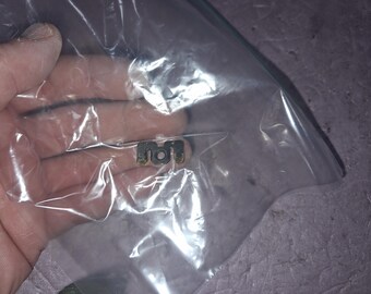 May include: A small, U-shaped metal component with a dark, weathered finish and a central circular hole, enclosed within a clear, crinkled plastic bag. The object features two distinct prongs and shows signs of oxidation or wear.