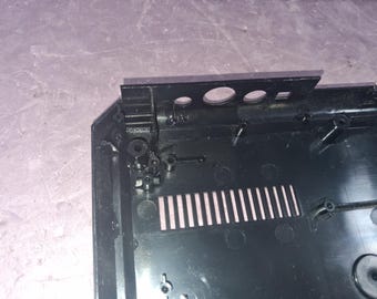 May include: A black plastic electronic device housing featuring a rectangular ventilation grille, internal mounting screw posts, and a top edge panel with three circular cutouts for ports or buttons, showing the interior structure.