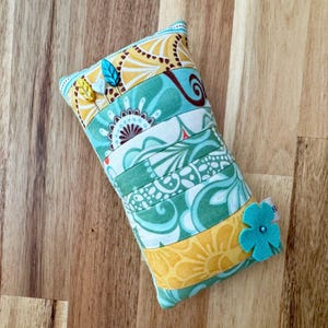 Pode incluir: Um almofada para alfinetes retangular com um design de patchwork em tecido amarelo, verde e branco. O almofada para alfinetes tem uma flor azul costurada em um lado e está cheio de alfinetes.