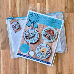 May include: A cross stitch pattern kit titled Homestead and Harmony featuring three round designs with a bird, a house, and a beehive. The kit includes a clear plastic storage pouch and a small circular tag with a bird motif, all displayed together.