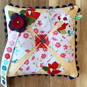 以下が含まれることがあります： 黄色と白の生地でできた、フリルのある縁取りと青いトリムが付いたピン刺し。ピン刺しは、赤と青のフェルトの花、白い布の花、緑の葉で飾られています。木製の頭をしたピンが2本、上から突き出ています。