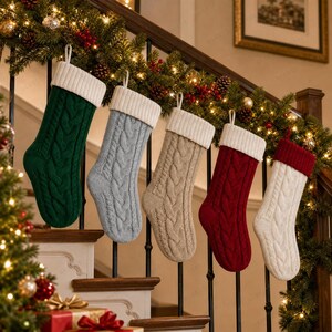 Op de afbeelding: Vijf kerstsokken met kabelpatroon hangend op een rij. De sokken zijn donkergroen, lichtgrijs, beige, dieprood en crème. Elke sok heeft een dikke, geribbelde witte boord, behalve de crèmekleurige sok die een rode boord heeft.