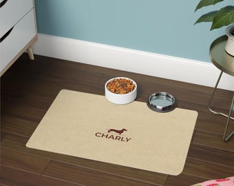 May include: A rectangular beige pet feeding mat featuring a brown silhouette of a dachshund dog above the text "CHARLY" in brown lettering. The mat is placed on a dark wood floor with a white bowl of dry food and an empty metal bowl.