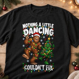 May include: A black short-sleeve t-shirt featuring a graphic of a brown Bigfoot wearing a Santa hat and a green Christmas tree dancing. The design includes colorful string lights, snowflakes, and the text "NOTHING A LITTLE DANCING COULDN'T FIX."