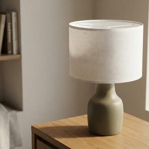 May include: A table lamp featuring a smooth, matte olive-green ceramic base with a rounded, tapered silhouette. The lamp is topped with a cylindrical, off-white fabric drum shade that provides a soft, diffused light effect.