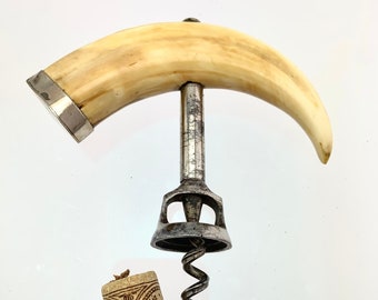 May include: A vintage corkscrew with a curved ivory handle and a metal screw. The corkscrew is resting on a white surface with a cork next to it.