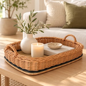 Peut inclure: Un plateau rectangulaire en osier tressé avec deux anses, contenant un vase en céramique blanche avec des brins verts et de petites fleurs blanches, une bougie pilier blanche allumée, un petit bol moucheté et un livre clair.