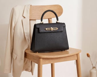 May include: A structured black handbag with a top handle and a gold-toned metal turn-lock closure. The bag features a flap design, a textured exterior finish, and four small gold-toned metal feet on the bottom base for stability.