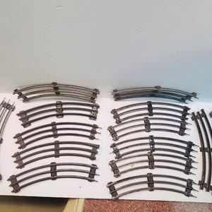 Op de afbeelding: Een verzameling van tweeëntwintig gebogen metalen modeltreinrails. Elk stuk is voorzien van drie parallelle rails verbonden door donkere metalen dwarsliggers. De rails zijn in meerdere rijen op een witte ondergrond gerangschikt.