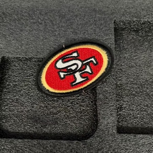 May include: An oval-shaped embroidered patch featuring the letters SF in white with a black outline. The letters are centered on a red background, which is surrounded by a gold border and a final black outer edge border.