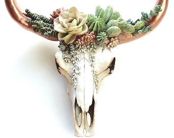 May include: A white ceramic bull skull with copper horns is decorated with a variety of green and pink succulents.
