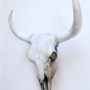 May include: A white painted bull skull with large, curved horns. The skull has visible cracks and a weathered appearance, with areas of brown and black paint. The horns are a bright white, contrasting with the skull's off-white color.