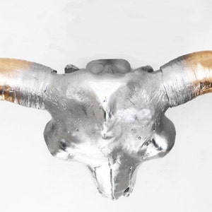 May include: A decorative bull skull with a silver finish and gold-colored horns. The skull is mounted on a white background. This piece is suitable for wall decor and adds a rustic touch to any space.