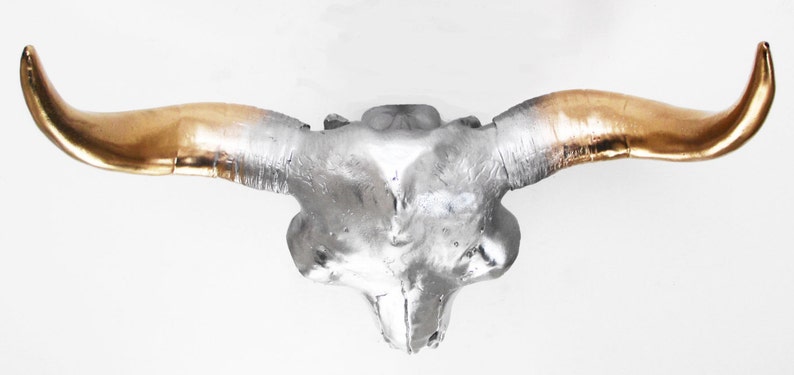 May include: A decorative bull skull with a silver finish and gold-colored horns. The skull is mounted on a white background. This piece is suitable for wall decor and adds a rustic touch to any space.