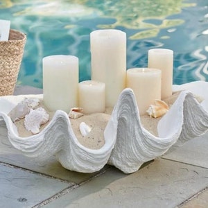May include: White ceramic clam shell with sand and seashells inside. Five white pillar candles are placed inside the shell.