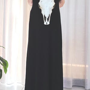 May include: A black sleeveless maxi dress featuring a white cow skull design. The skull has black horns and is held in front of the dress. The dress is floor-length and the background is a white curtain.