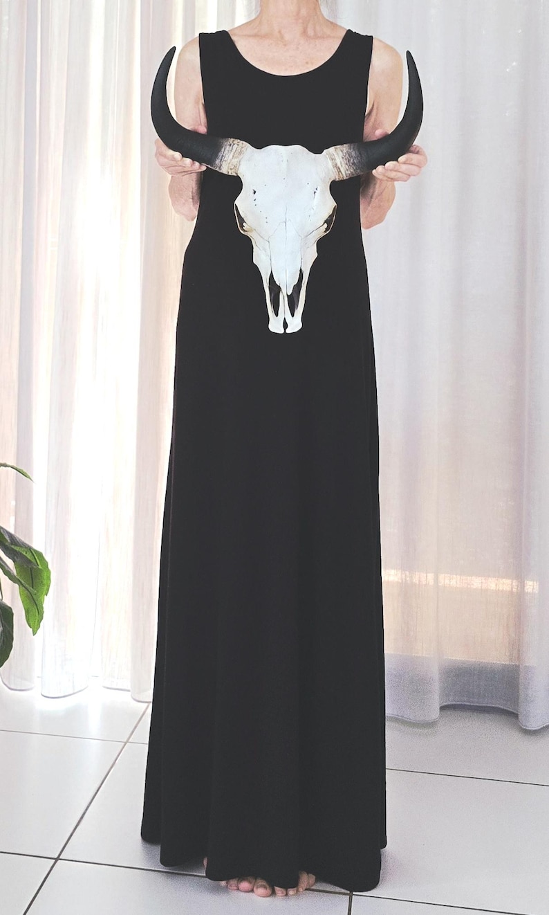May include: A black sleeveless maxi dress featuring a white cow skull design. The skull has black horns and is held in front of the dress. The dress is floor-length and the background is a white curtain.
