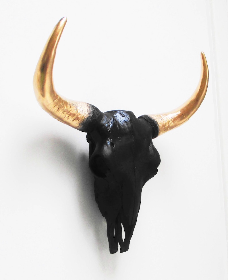 May include: A black bull skull wall decor with gold-colored horns. The skull is painted black, and the horns curve upwards. This decorative piece is designed to be mounted on a wall.