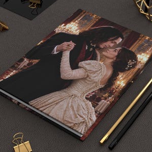 May include: A rectangular hardcover journal featuring a printed image of a couple dancing in a ballroom. The person on the left wears a black suit with a red vest, while the person on the right wears a cream-colored gown with long sleeves.