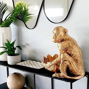 Op de afbeelding: Een gouden apenbeeld staat op een zwart rek met een zwart-wit gepatroneerd dienblad. Het rek maakt deel uit van een zwart metalen rek. Twee golvende spiegels met zwarte frames hangen aan de muur achter het rek.