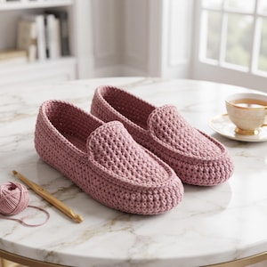 May include: A pair of dusty rose crochet loafers with a textured, hand-stitched appearance. The shoes feature a rounded toe and a slip-on design. A small ball of matching yarn and a wooden crochet hook are positioned next to the footwear.