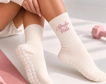 Puede incluir: Un par de calcetines de corte medio acanalados en color crema con el texto Studio Vela bordado en rosa en el tobillo exterior. Las suelas de los calcetines tienen un patrón de pequeños puntos de agarre de silicona blanca en forma de corazón.