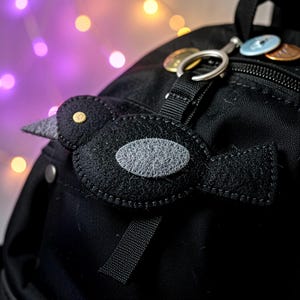 May include: A black felt bird ornament with a grey wing patch and a yellow eye detail, attached to a black backpack by a silver metal ring. The bird features visible stitching along its edges and is positioned on the front of the bag.