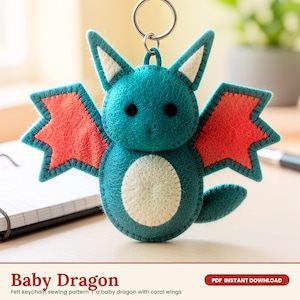 May include: A teal felt keychain shaped like a baby dragon with coral-colored wings, cream-colored inner ears, and a cream-colored belly. The creature has two small black eyes and is attached to a silver metal keyring at the top.
