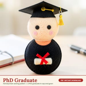 May include: A felt keychain shaped like a graduate wearing a black cap and gown. The figure has a beige head with black eyes and pink cheeks, a black body, and a small white diploma tied with a red ribbon. A yellow tassel hangs from the cap.