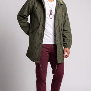 May include: An olive green hooded parka with a dark green grid pattern, worn over a white V-neck t-shirt and maroon trousers. The outfit is completed with a silver chain necklace featuring two rectangular pendants and olive and white sneakers.