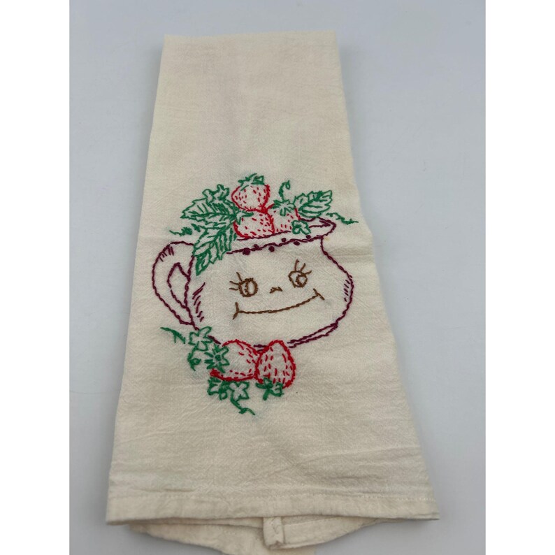 May include: Cream-colored kitchen towel with an embroidered design of a smiling pitcher filled with strawberries. The pitcher is outlined in dark red thread, with green leaves and red strawberries. The towel is folded.
