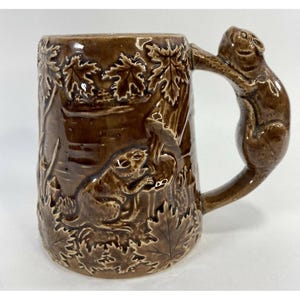 May include: Brown ceramic mug with a detailed relief of squirrels, leaves, and trees. The handle is shaped like a squirrel. The mug has a glossy finish and is suitable for hot or cold beverages.