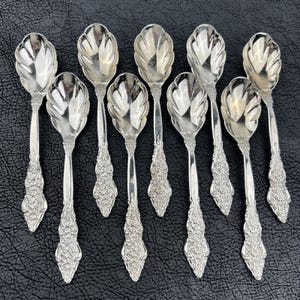May include: A set of eight silver-toned spoons featuring a polished bowl with a scalloped shell-like edge and a handle decorated with intricate floral and scroll relief patterns. The spoons are arranged in two rows against a black textured surface.