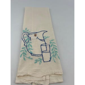 May include: Cream-colored kitchen towel with an embroidered design. The design features a blue pitcher, a glass, and leafy sprigs. The embroidery is in shades of blue and the towel is folded.