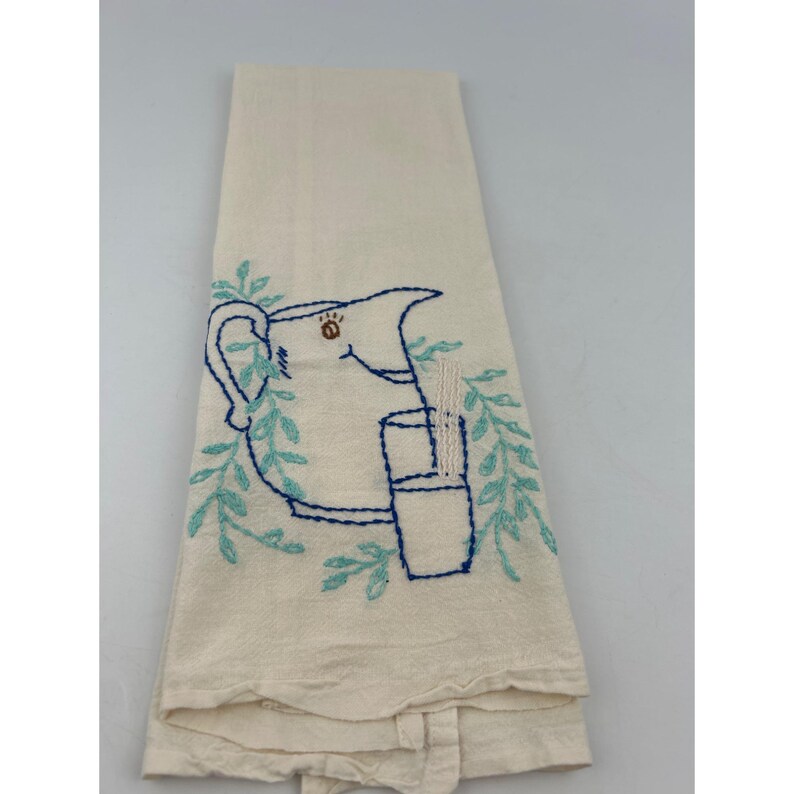 May include: Cream-colored kitchen towel with an embroidered design. The design features a blue pitcher, a glass, and leafy sprigs. The embroidery is in shades of blue and the towel is folded.