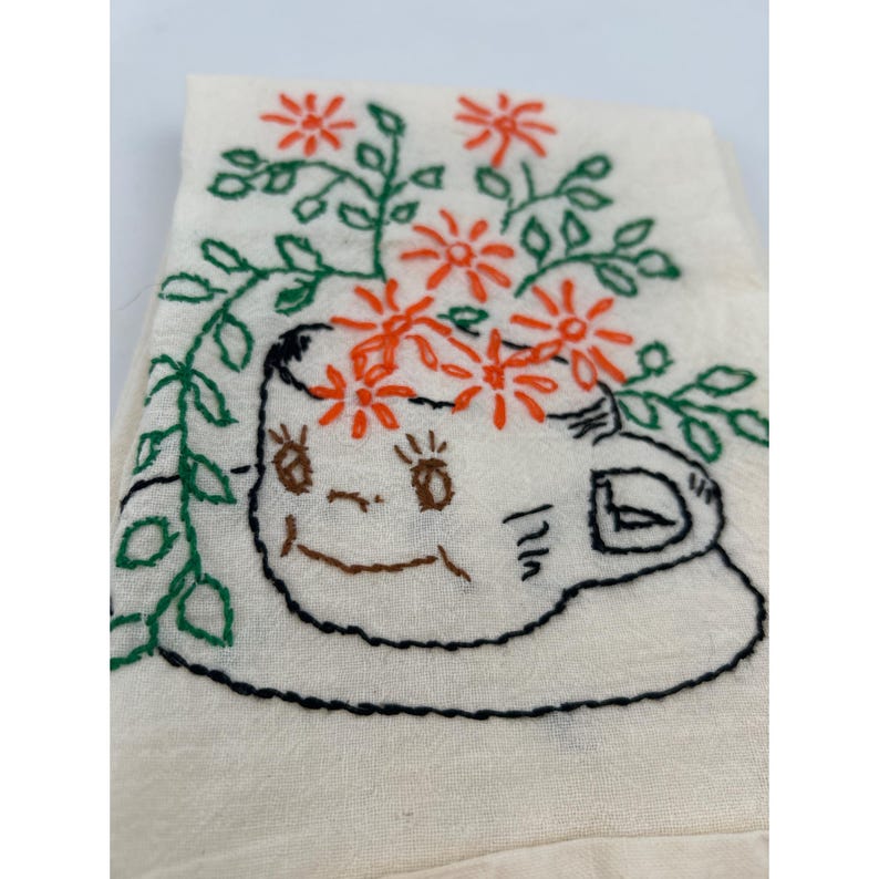 May include: Embroidered off-white tea towel featuring a whimsical teacup design. The teacup has a smiling face and is adorned with orange flowers and green leaves. The design is outlined in black and brown thread.