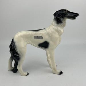 May include: A ceramic Borzoi dog figurine in a standing pose. The dog is primarily white with black markings on its head, back, and legs. The figurine has a glossy finish and a small label that reads "Duke".