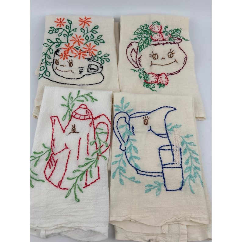 May include: Four off-white embroidered kitchen towels. Each towel features a different design: a teacup with flowers, a teapot with strawberries, a red teapot, and a pitcher with a glass. The embroidery uses red, green, blue, and black thread.