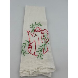 May include: White tea towel with a red embroidered teapot design and green leaf accents. The tea towel is folded and has a textured appearance. A charming kitchen textile.
