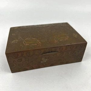 May include: A rectangular, antique-style wooden box with a hinged lid. The box is a warm brown color and features an intricate floral and leaf pattern carved into the surface. The box has a small metal clasp.