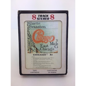 May include: Vintage 8-track stereo tape cartridge for the band Chicago. The label features the band's logo in orange and the names of cities like Wilmette, Evanston, and East Chicago. The cartridge is black with white text.
