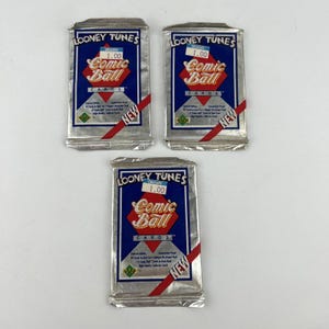 May include: Three sealed packs of Looney Tunes Comic Ball cards. Each pack has a silver foil wrapper with a blue and red design, featuring the text "Comic Ball Cards" and the Looney Tunes logo. The packs are labeled "NEW".
