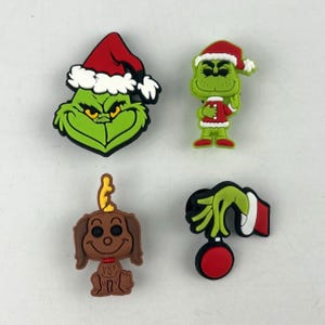 May include: A set of four shoe charms featuring a green character with a red Santa hat, a full-body green character in a Santa suit, a brown dog with long ears and a yellow antler, and a green hand wearing a red sleeve holding a red ornament.