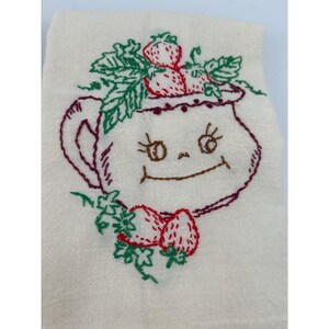May include: Cream-colored embroidered tea towel featuring a smiling strawberry pot design. The pot is outlined in burgundy with a smiling face, and topped with green leaves and red strawberries. Additional strawberries and leaves are embroidered below.