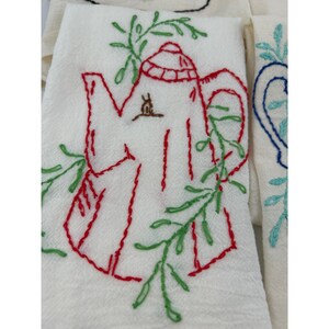 May include: White tea towel with a red embroidered teapot design, accented with green and red leaves. The teapot has a red lid and handle. Additional towels with blue and black embroidered designs are visible.