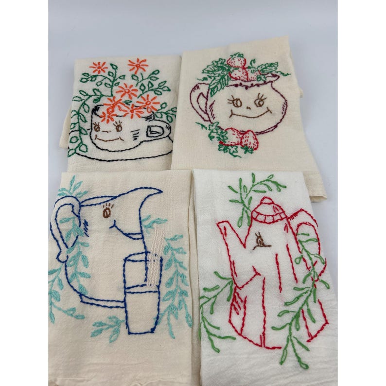 May include: Four off-white embroidered kitchen towels. Each towel features a different whimsical design: a teacup with flowers, a teapot with strawberries, a pitcher with a glass, and a teapot with leaves. The embroidery uses red, blue, and green thread.